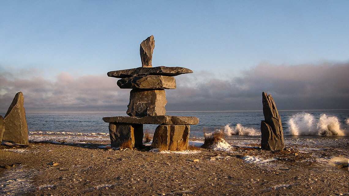 Stone Inukshuk, or traditional Inuit stone wayfinding marker on gravel beach. Small drifts of snow at base of marker and waves crashing against the shoreline behind Inukshuk. The sky is blue with storm clouds in the distance near the horizon.